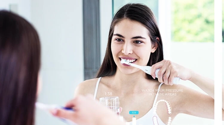 A joyful girl brushes her teeth and holds a glass of water while looking into the mirror.